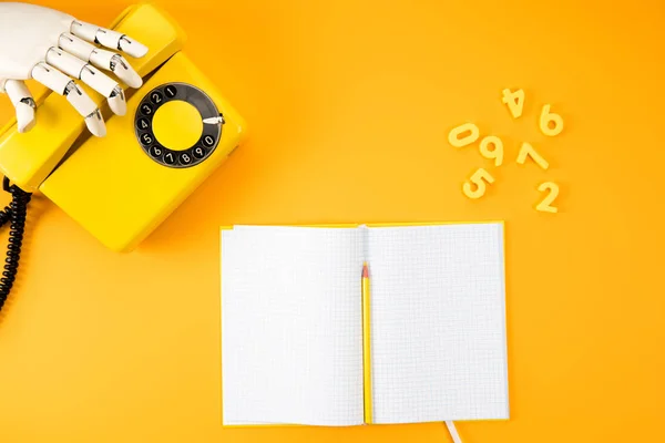 Tiro recortado da mão robótica que alcança para o telefone do vintage na mesa amarela com notebook em branco e números de matemática — Fotografia de Stock