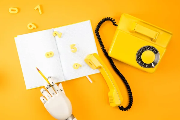 Cropped shot of robotic hand writing in blank notebook on yellow tabletop with vintage phone and math numbers — Stock Photo