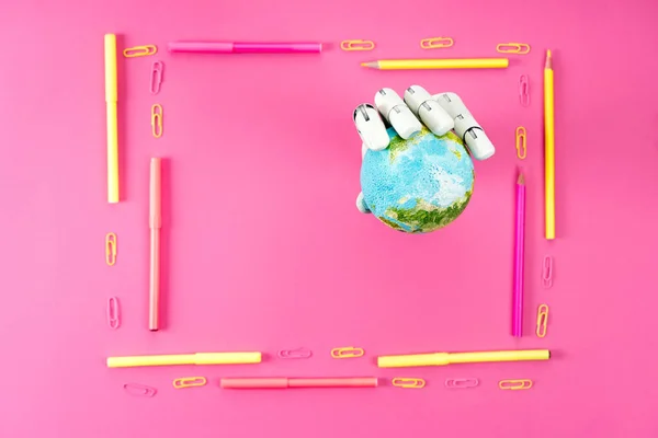 Top view of robotic hand holding globe inside of frame made of pencils and markers on pink — Stock Photo