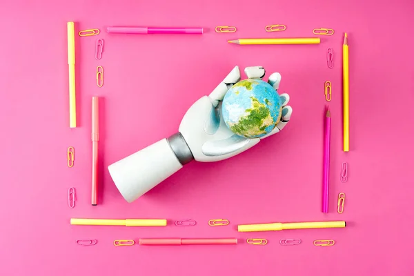 Top view of robotic hand holding earth globe inside of frame made of pencils and markers on pink — Stock Photo