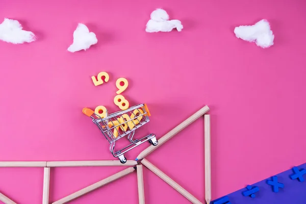 Shopping cart with various numbers riding on cliff arranged with color pencils to mathematical expression on pink — Stock Photo