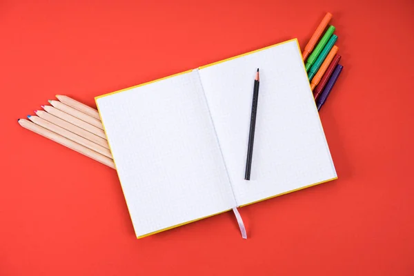 Vue du haut du bloc-notes vierge ouvert avec crayons de couleur et marqueurs sur rouge — Photo de stock