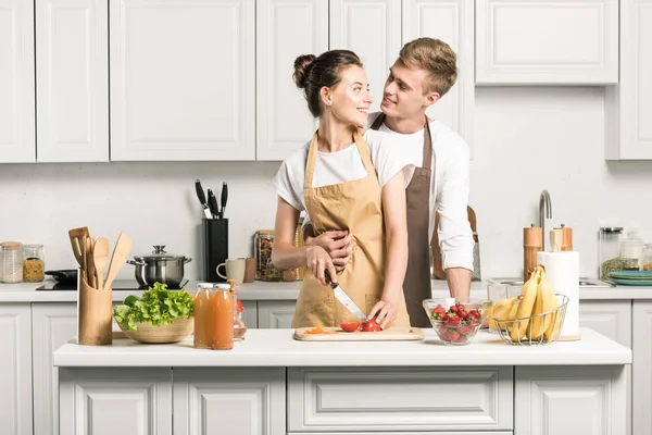 Парень обнимает девушку, пока она готовит салат на кухне — стоковое фото