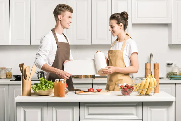 Joven pareja cocina ensalada y celebración de papel toalla en la cocina - foto de stock