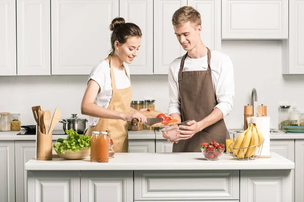 Jovem casal cozinhar salada e colocar legumes na tigela na cozinha — Fotografia de Stock