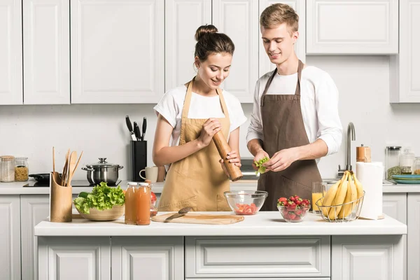 Pareja joven cocinar ensalada y añadir especias en la cocina - foto de stock