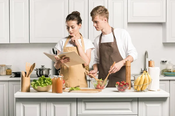 Young couple cooking salad and reading recipe in cookbook in kitchen — Stock Photo