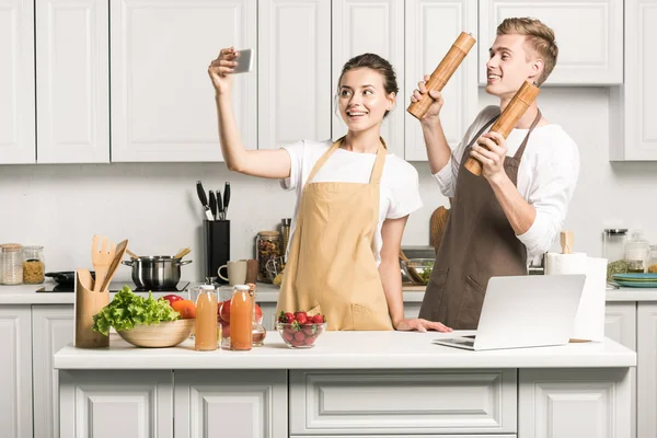 Молодая пара делает селфи со смартфоном во время приготовления пищи на кухне — стоковое фото