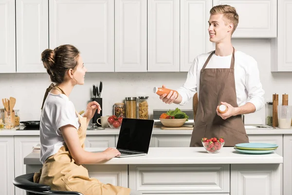 Girlfriend using laptop, boyfriend holding bottles of juice in kitchen — Stock Photo