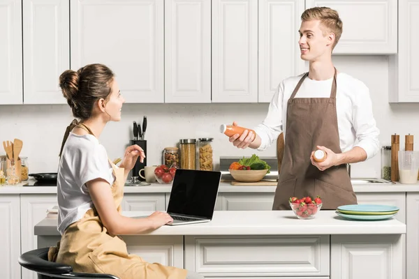 Novia usando portátil, novio sosteniendo botellas de jugo en la cocina - foto de stock