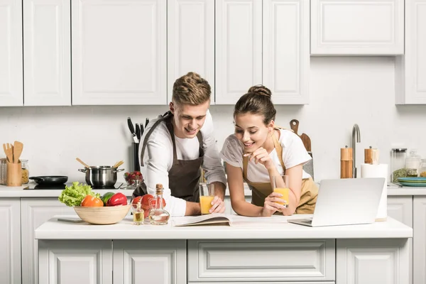 Jeune couple lisant le livre de recettes et tenant des verres avec du jus sain dans la cuisine — Photo de stock