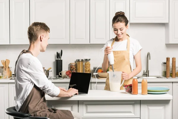 Petit ami utilisant un ordinateur portable, petite amie préparant milkshake dans la cuisine — Photo de stock