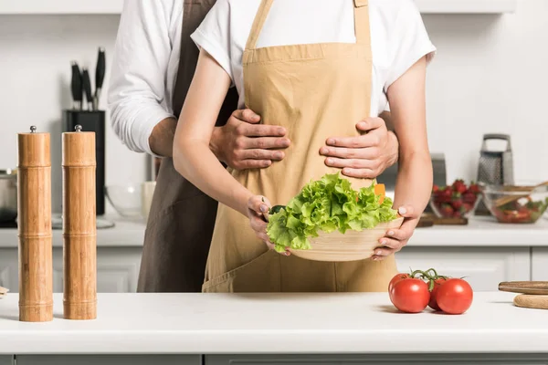 Обрізане зображення молодої пари, що обіймає і тримає миску з салатом на кухні — стокове фото