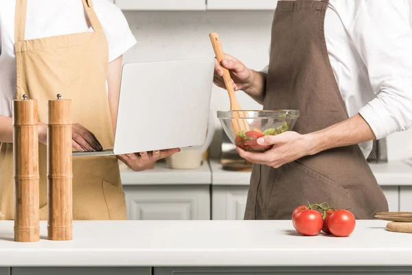 Imagen recortada de pareja de ensalada de cocina y el uso de ordenador portátil en la cocina - foto de stock