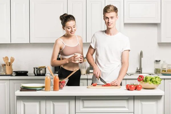 Jovem casal cozinhar salada, namorado cortar tomates na cozinha — Fotografia de Stock
