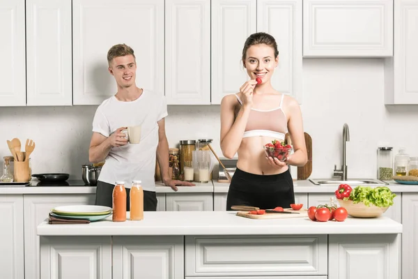 Girlfriend in sport bra eating strawberry in kitchen — Stock Photo