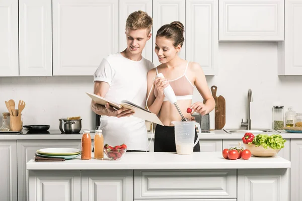 Giovane coppia che prepara frullati con ricettario in cucina — Foto stock