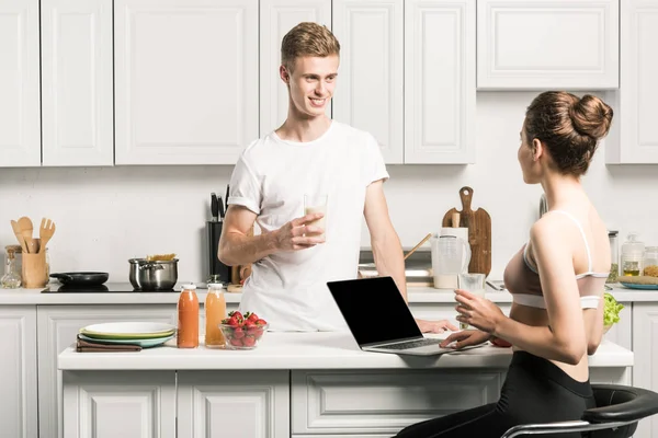 Petit ami tenant verre de lait et petite amie en utilisant un ordinateur portable dans la cuisine — Photo de stock