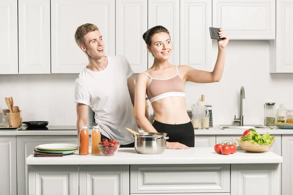Дівчина в спортивному бюстгальтері бере селфі зі смартфоном на кухні — стокове фото