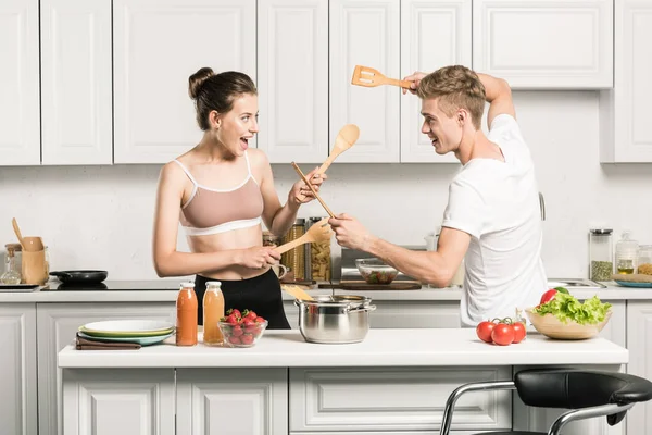 Молодая пара, дерущаяся с деревянными лопатками на кухне — стоковое фото