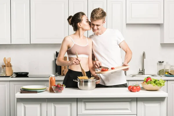 Petite amie embrasser petit ami tout en cuisinant des pâtes dans la cuisine — Photo de stock