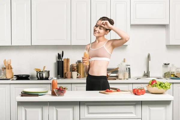 Attractive girl in sport bra touching forehead and holding bottle of fresh juice in kitchen — Stock Photo