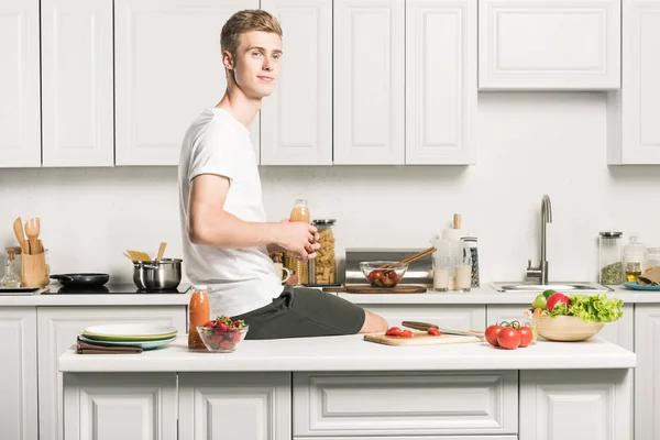 Handsome young man sitting on kitchen counter and holding healthy juice in bottle — Stock Photo