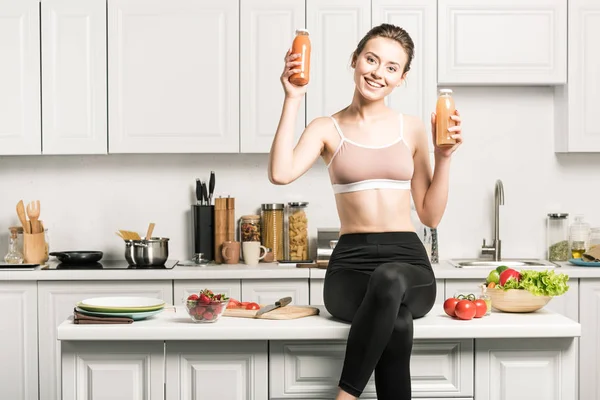 Happy attractive girl sitting on kitchen counter and showing healthy juice in bottles — Stock Photo