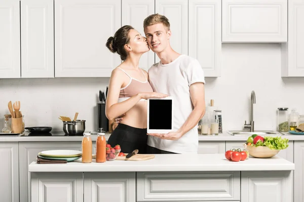 Namorada beijando namorado e ele segurando tablet com tela em branco na cozinha — Fotografia de Stock