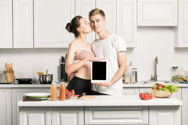 Fidanzata baciare fidanzato e lui tenendo tablet con schermo bianco in cucina — Foto stock