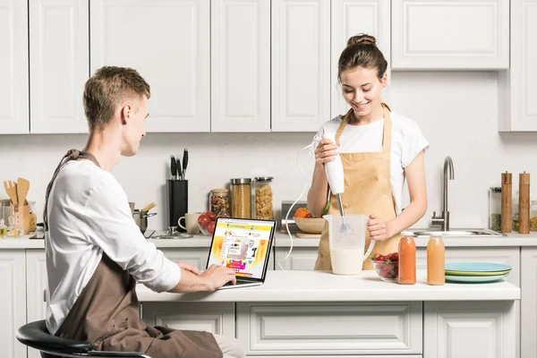 Fidanzata cucina e fidanzato utilizzando computer portatile con caricato aliexpress pagina in cucina — Foto stock