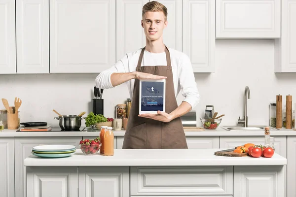 Bellissimo uomo in grembiule mostrando tablet con pagina tumblr caricato in cucina — Foto stock