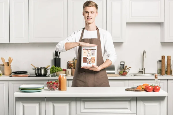 Bonito homem no avental mostrando tablet com carregado pinterest página na cozinha — Fotografia de Stock