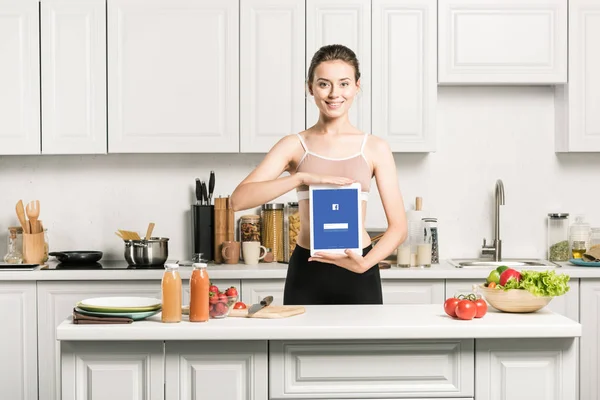 Atractiva chica sosteniendo tableta con cargado facebook página en la cocina - foto de stock