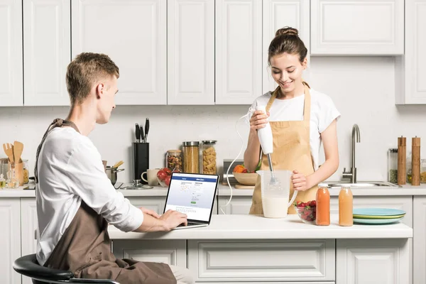 Namorada cozinhar e namorado usando laptop com página Facebook carregada na cozinha — Fotografia de Stock