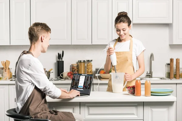 Novia cocina y novio usando el ordenador portátil con página linkedin cargado en la cocina - foto de stock