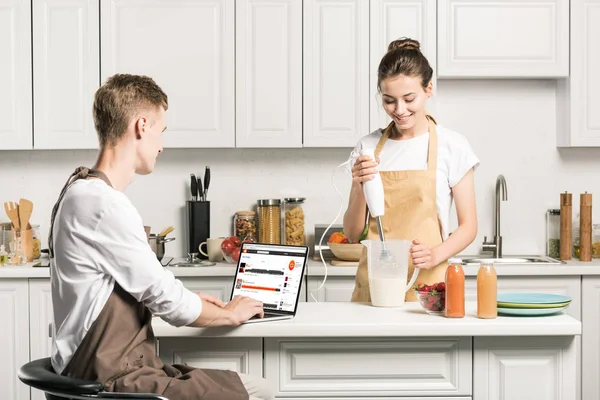 Novia cocina y novio usando el ordenador portátil con la página de soundcloud cargado en la cocina - foto de stock