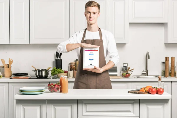 Bel homme dans tablier montrant tablette avec page instagram chargé dans la cuisine — Photo de stock