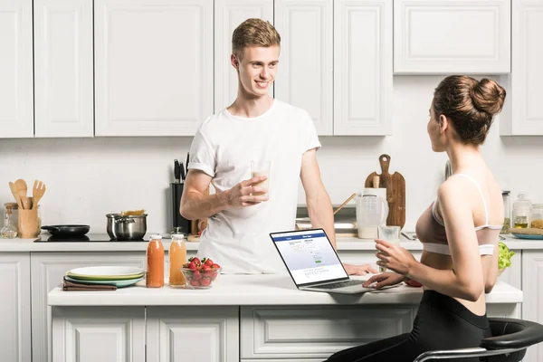 Girlfriend using laptop with loaded facebook page and looking at boyfriend in kitchen — Stock Photo