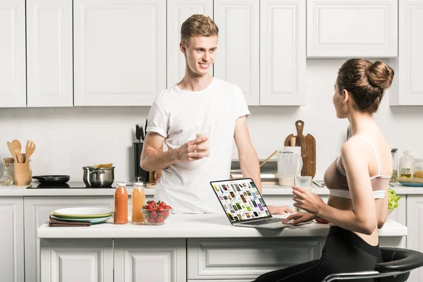 Girlfriend using laptop with loaded pinterest page and looking at boyfriend in kitchen — Stock Photo