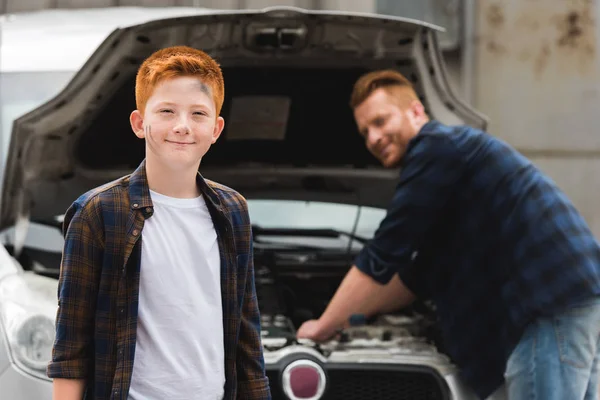 Padre reparación de coche con capucha abierta, sonriente hijo mirando a la cámara - foto de stock