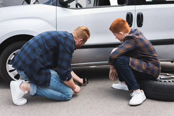 Father lifting car with floor jack for changing tire, son sitting on tire — Stock Photo