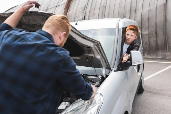 Father repairing car with open hood, son sitting at driver seat — Stock Photo