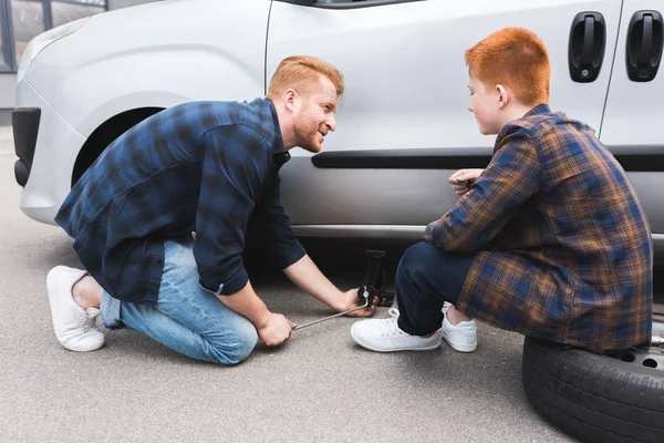 Padre levantando coche con gato de piso para cambiar el neumático, hijo mirándolo - foto de stock