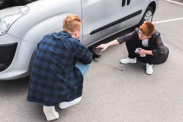 Padre levantando coche con gato de piso para cambiar el neumático, hijo señalando algo - foto de stock