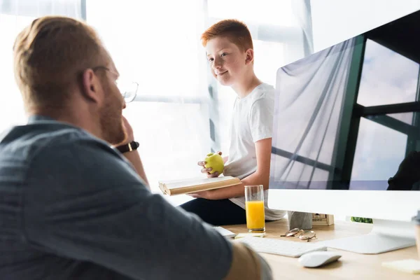 Hijo sosteniendo libro y manzana y mirando a padre en casa - foto de stock