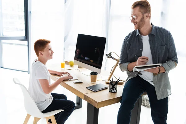 Father holding pen and notebook, son using computer at home — Stock Photo