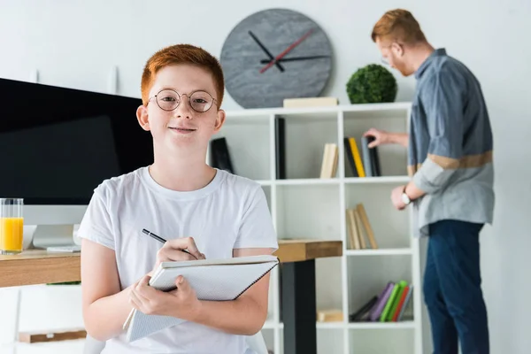 Ginger hair boy holding pen and notebook and looking at camera at home — Stock Photo