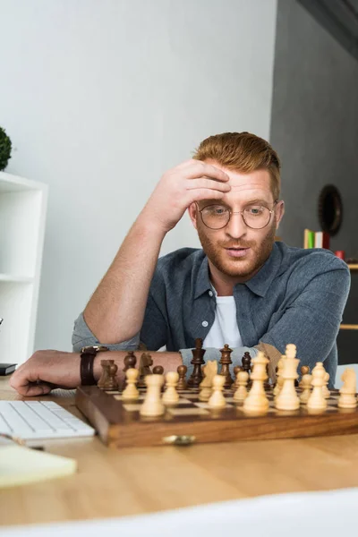 Bonito homem pensativo olhando para tabuleiro de xadrez em casa — Fotografia de Stock