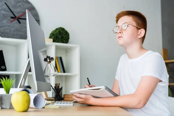 Pensive preteen ginger hair boy holding pen with notebook at home and looking away — Stock Photo